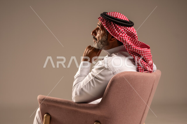 التأمل وشرود الذهن، التفكير في شيء هام، بورتريه من الجانب لرجل مسن عربي خليجي سعودي يرتدي الثوب التقليدي والشماغ يجلس على الكرسي يضع يده على ذقنه وينظر الى شيء، خلفية بيج