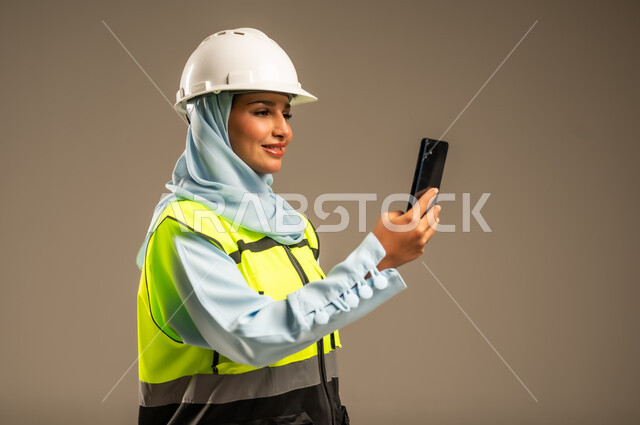 Searching for engineering plans via mobile, integrating work with technology and technology, close-up portrait of a Saudi Arabian Gulf female architect wearing a helmet and a protective vest using a mobile phone, female engineering professions and jobs, using technology in engineering fields, gray background