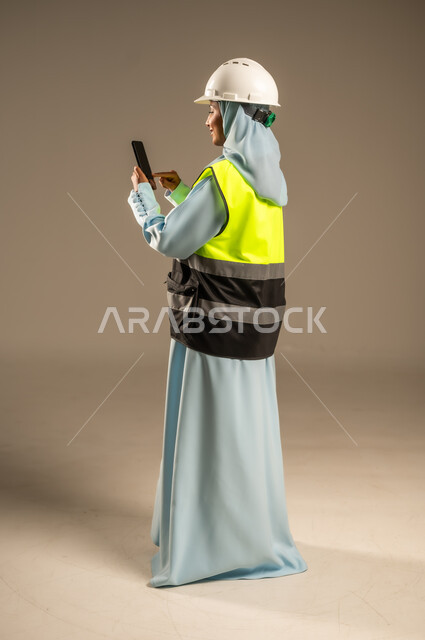 Integrating work with technology and technology, searching for engineering drawings via mobile, portrait from the back of a Saudi Arabian Gulf female engineer wearing a helmet and jacket using a mobile phone, female engineering professions and jobs, using technology in engineering fields, full-length body, gray background
