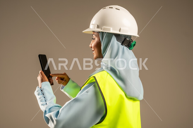 البحث عن المخططات الهندسيه عبر الجوال, دمج العمل مع التقنية و التكنولوجيا, بورتريه مقرب من الخلف لمهندسة معمارية عربية خليجية سعودية ترتدي الخوذة وسترة الحماية تستخدم الهاتف المحمول، مهن ووظائف هندسية نسائية, استخدام التقنيات في المجالات الهندسية, خلفية رمادية