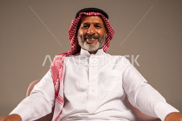 تعابير البشاشة والسرور، الاهتمام بالمظهر الخارجي، بورتريه مقرب لرجل كبير بالسن عربي خليجي سعودي يرتدي الشماغ والثوب التقليدي يجلس على كرسي وينظر الى الكاميرا، خلفية بيج
