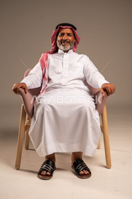 ايماءات البشاشة والسرور، الاهتمام بالمظهر الخارجي، بورتريه لرجل كبير بالسن عربي خليجي سعودي يرتدي الشماغ والثوب التقليدي يجلس على كرسي وينظر الى الكاميرا، خلفية بيج