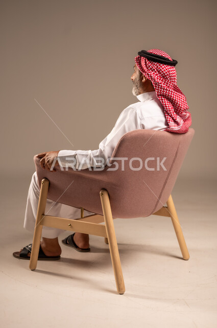 التأمل وشرود الذهن، التفكير في شيء هام، بورتريه من الجانب لرجل مسن عربي خليجي سعودي يرتدي الثوب التقليدي والشماغ يجلس على الكرسي ينظر الى شيء، خلفية بيج