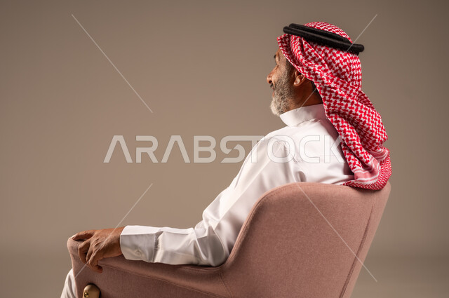 التأمل وشرود الذهن، التفكير في شيء هام، بورتريه من الجانب لرجل مسن عربي خليجي سعودي يرتدي الثوب التقليدي والشماغ يجلس على الكرسي ينظر الى شيء، خلفية بيج