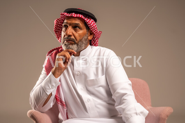 النظر إلى شيء ما، التأمل والتخطيط للمستقبل، الاهتمام بالمظهر الخارجي، بورتريه مقرب لرجل مسن عربي خليجي سعودي يرتدي الثوب التقليدي والشماغ يجلس على كرسي مريح ويضع يده تحت ذقنه، خلفية بيج