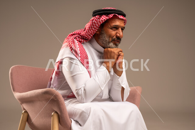 الاهتمام بالمظهر الخارجي، بورتريه مقرب لرجل مسن عربي خليجي سعودي يرتدي الثوب التقليدي والشماغ يجلس على كرسي مريح ويضع يده تحت ذقنه، خلفية بيج