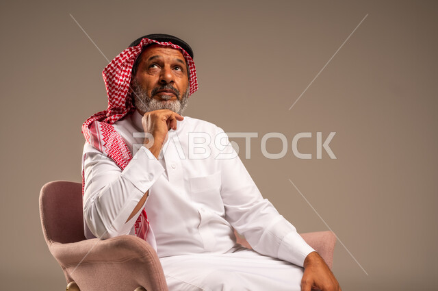 النظر إلى شيء ما، التأمل والتخطيط للمستقبل، الاهتمام بالمظهر الخارجي، بورتريه مقرب لرجل مسن عربي خليجي سعودي يرتدي الثوب التقليدي والشماغ يجلس على كرسي مريح ويضع يده تحت ذقنه، خلفية بيج