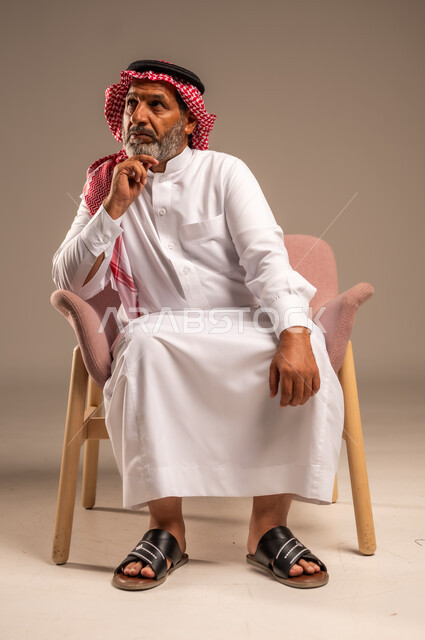 الاهتمام بالمظهر الخارجي، التأمل والتخطيط للمستقبل، بورتريه لرجل مسن عربي خليجي سعودي يرتدي الثوب التقليدي والشماغ يجلس على كرسي مريح ويضع يده تحت ذقنه، خلفية بيج