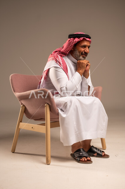 الاهتمام بالمظهر الخارجي، التأمل والتخطيط للمستقبل، بورتريه لرجل مسن عربي خليجي سعودي يرتدي الثوب التقليدي والشماغ يجلس على كرسي مريح ويضع يده تحت ذقنه، خلفية بيج