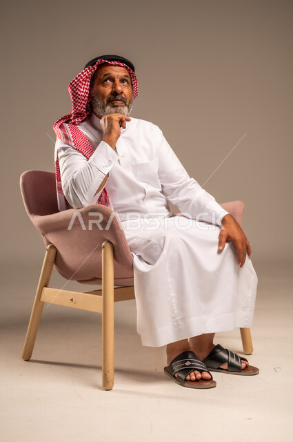 الاهتمام بالمظهر الخارجي، التأمل والتخطيط للمستقبل، بورتريه لرجل مسن عربي خليجي سعودي يرتدي الثوب التقليدي والشماغ يجلس على كرسي مريح ويضع يده تحت ذقنه، خلفية بيج