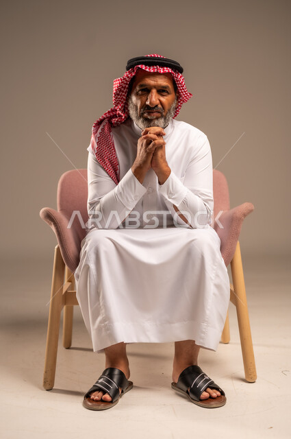 الاهتمام بالمظهر الخارجي، بورتريه لرجل كبير بالسن عربي خليجي سعودي يرتدي الثوب التقليدي والشماغ يجلس على كرسي مريح ويضع يده تحت ذقنه ينظر الى الكاميرا بايماءات السرور، خلفية بيج