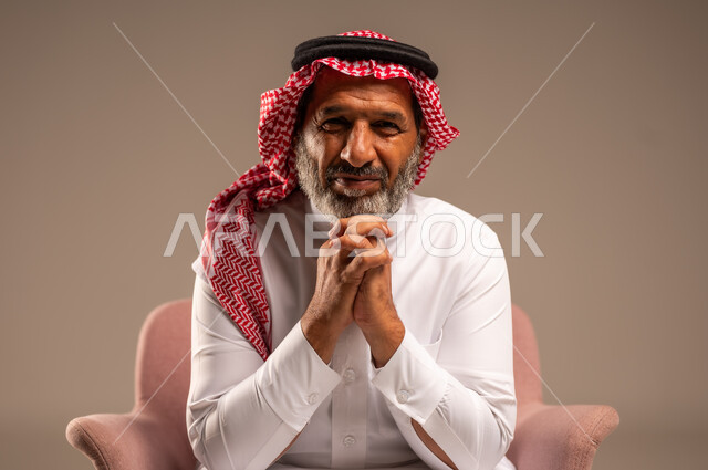 الاهتمام بالمظهر الخارجي، بورتريه مقرب لرجل كبير بالسن عربي خليجي سعودي يرتدي الثوب التقليدي والشماغ يجلس على كرسي مريح ويضع يده تحت ذقنه ينظر الى الكاميرا بايماءات السرور، خلفية بيج
