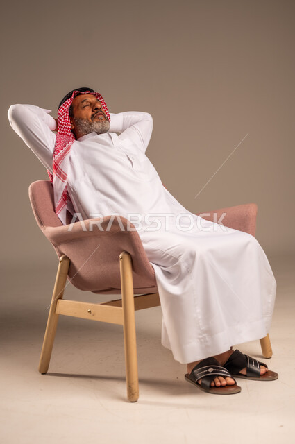 الاستمتاع بوقت الراحة والهدوء, أخذ قسط من الاسترخاء و التأمل, بورتريه من الجانب لرجل مسن عربي خليجي سعودي يرتدي الثوب التقليدي والشماغ يجلس على كرسي مريح, خلفية بيج