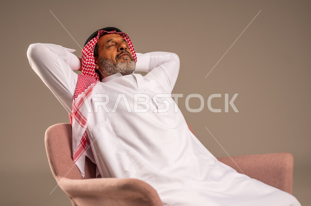 ايماءات التأمل والاستمتاع بوقت الهدوء، أخذ قسط من الراحة، بورتريه مقرب لرجل كبير بالسن يرتدي الثوب التقليدي والشماغ يجلس على كرسي يضع يديه خلف رأسه بتعابير الاسترخاء والسرور، خلفية بيج