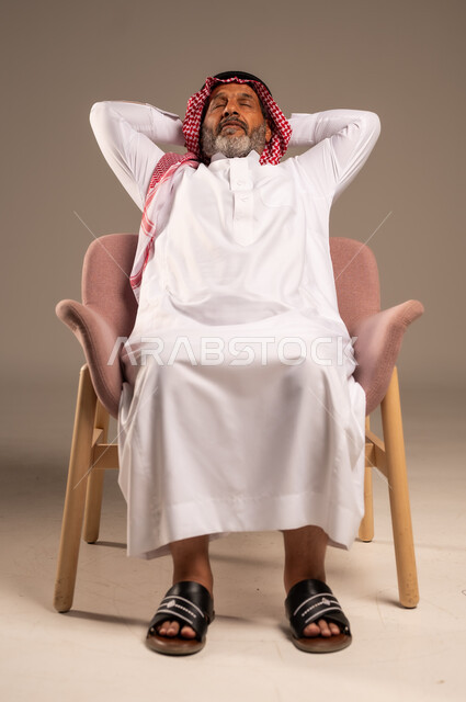 أخذ قسط من الاسترخاء و التأمل، الاستمتاع بوقت الراحة والهدوء, بورتريه لرجل مسن عربي خليجي سعودي يرتدي الثوب التقليدي والشماغ يجلس على كرسي مريح ينظر الى شيء ما بتعابير السرور, خلفية بيج