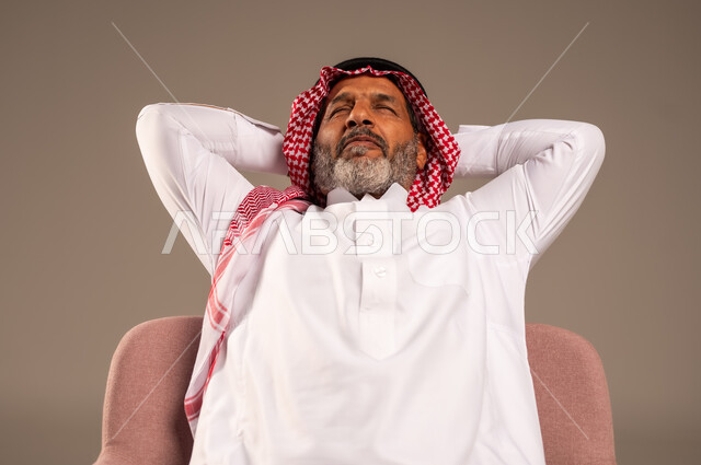 أخذ قسط من الاسترخاء و التأمل، الاستمتاع بوقت الراحة والهدوء, بورتريه لرجل مسن عربي خليجي سعودي يرتدي الثوب التقليدي والشماغ يجلس على كرسي مريح ينظر الى شيء ما بتعابير السرور, خلفية بيج