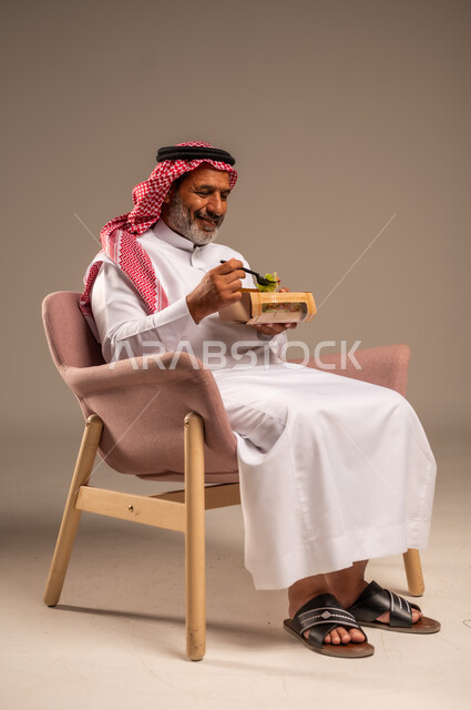 توفير العناصر الغذائية الضرورية للجسم، تناول وجبات الطعام الصحية، بورتريه لرجل مسن عربي خليجي سعودي يرتدي زي تقليدي وشماغ يجلس على كرسي يحمل بيده طبق، الحفاظ على الرشاقة والصحة البدنية، خلفية بيج