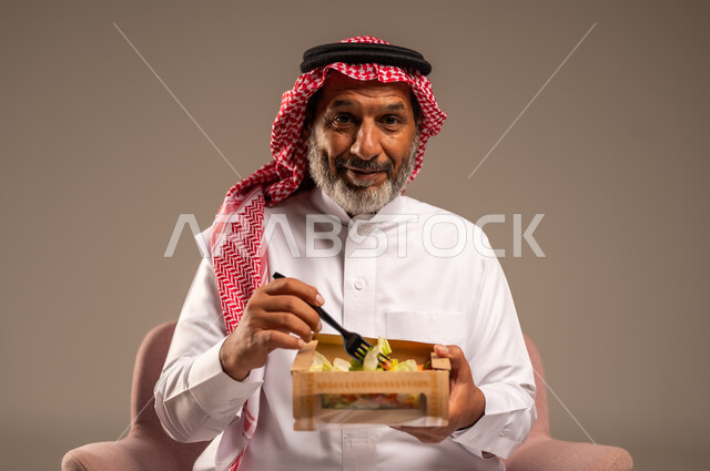 الحفاظ على الرشاقة والصحة البدنية، توفير العناصر الغذائية الضرورية للجسم، تناول وجبات الطعام الصحية، بورتريه لرجل مسن عربي خليجي سعودي يرتدي زي تقليدي وشماغ يجلس على كرسي يحمل بيده طبق من السلطة، النظر الى الكاميرا بايماءات السرور، خلفية بيج