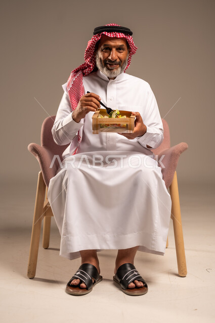 الحفاظ على الرشاقة والصحة البدنية، توفير العناصر الغذائية الضرورية للجسم، تناول وجبات الطعام الصحية، بورتريه لرجل مسن عربي خليجي سعودي يرتدي زي تقليدي وشماغ يجلس على كرسي يحمل بيده طبق ينظر الى الكاميرا بايماءات السرور، خلفية بيج