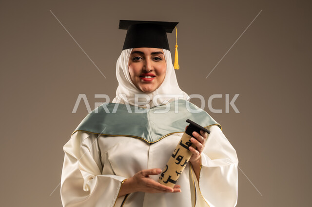 الاحتفال بإنهاء الدراسة الجامعية، ايماءات البهجة والسعادة بالوصول لحلم النجاح, بورتريه مقرب لخريجة عربية سعودية خليجية محجبة ترتدي العباءة البيضاء وقبعة التخرج تمسك بيدها وثيقة التخرج تنظر الى الكاميرا, خلفية بيج