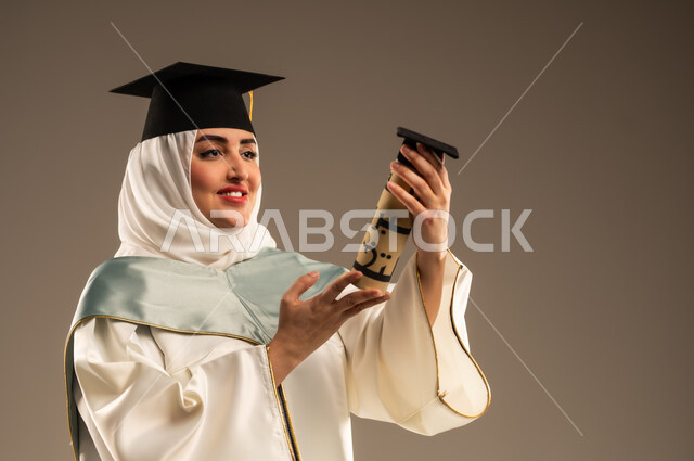 الاحتفال بإنهاء الدراسة الجامعية، ايماءات البهجة والسعادة بالوصول لحلم النجاح, بورتريه مقرب لخريجة عربية سعودية خليجية محجبة ترتدي العباءة البيضاء وقبعة التخرج تمسك بيدها وثيقة التخرج, خلفية بيج