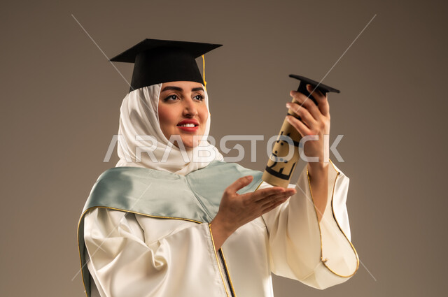 الاحتفال بإنهاء الدراسة الجامعية، ايماءات البهجة والسعادة بالوصول لحلم النجاح, بورتريه مقرب لخريجة عربية سعودية خليجية محجبة ترتدي العباءة البيضاء وقبعة التخرج تمسك بيدها وثيقة التخرج, خلفية بيج