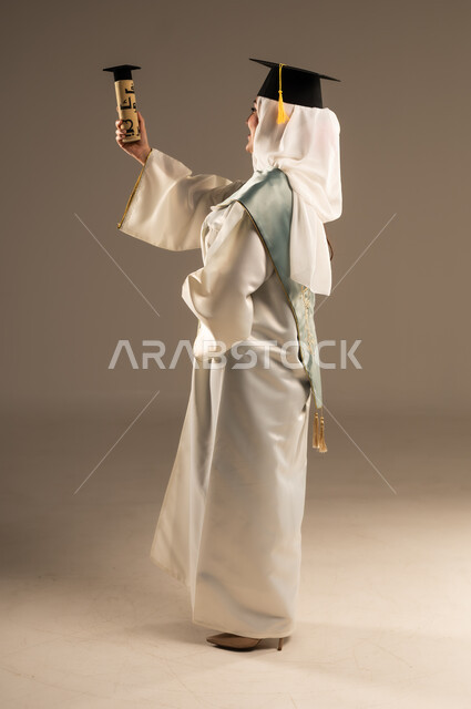 Celebrating the completion of university studies, gestures of joy and happiness at reaching the dream of success, portrait of a veiled Saudi Arabian Gulf graduate wearing a white abaya and graduation cap holding her graduation document in her hand, full-length body image, beige background