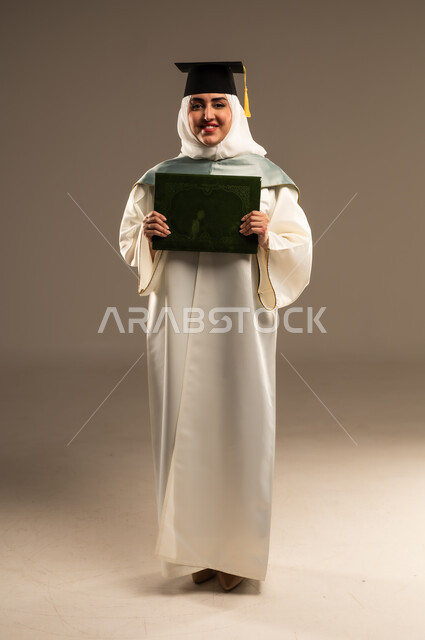 الاحتفال بإنهاء الدراسة الجامعية، ايماءات البهجة والسعادة بالوصول لحلم النجاح, بورتريه لطالبة عربية سعودية خليجية محجبة ترتدي العباءة البيضاء وقبعة التخرج تمسك بيدها شهادة التفوق, صورة الجسم بالطول الكامل, خلفية بيج
