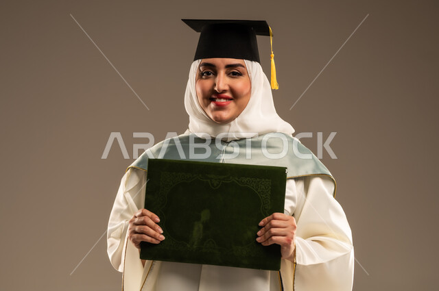الاحتفال بإنهاء الدراسة الجامعية، ايماءات البهجة والسعادة بالوصول لحلم النجاح, بورتريه مقرب لخريجة عربية سعودية خليجية محجبة ترتدي العباءة البيضاء وقبعة التخرج تمسك بيدها وثيقة التخرج تنظر الى الكاميرا, خلفية بيج