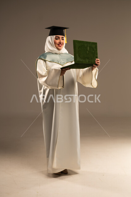 الاحتفال بتحقيق النجاح, أهمية الدراسة الجامعية, ايماءات البهجة والسعادة بالوصول لحلم التخرج، بورتريه لخريجة عربية سعودية خليجية محجبة ترتدي العباءة البيضاء وقبعة التخرج تنظر الى شيء ما تمسك بيدها شهادة التفوق, صورة الجسم بالطول الكامل, خلفية بيج