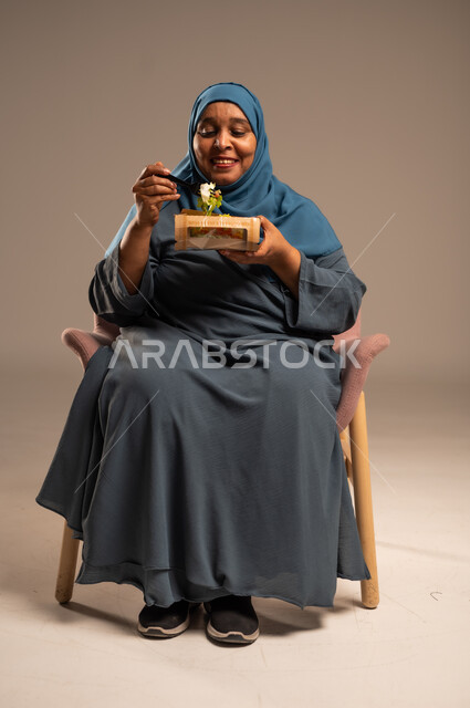 توفير العناصر الغذائية الضرورية للجسم، بورتريه لامراة مسنة عربية خليجية سعودية محجبة ترتدي العباءة الزرقاء تجلس على كرسي مريح تقوم بتناول وجبات الطعام الصحية، الحفاظ على الرشاقة والصحة البدنية، خلفية بيج