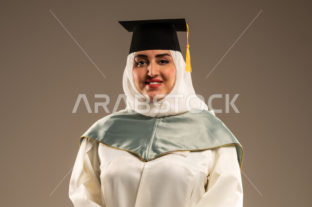 ايماءات البهجة والسعادة بالوصول لحلم النجاح, الاحتفال بإنهاء الدراسة الجامعية، بورتريه مقرب لخريجة عربية سعودية خليجية محجبة ترتدي العباءة البيضاء وقبعه التخرج تنظر الى الكاميرا, خلفية بيج