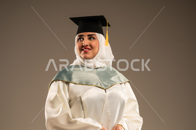 ايماءات البهجة والسعادة بالوصول لحلم النجاح, الاحتفال بإنهاء الدراسة الجامعية، بورتريه مقرب لخريجة عربية سعودية خليجية محجبة ترتدي العباءة البيضاء وقبعه التخرج تنظر الى شيئ ما, خلفية بيج