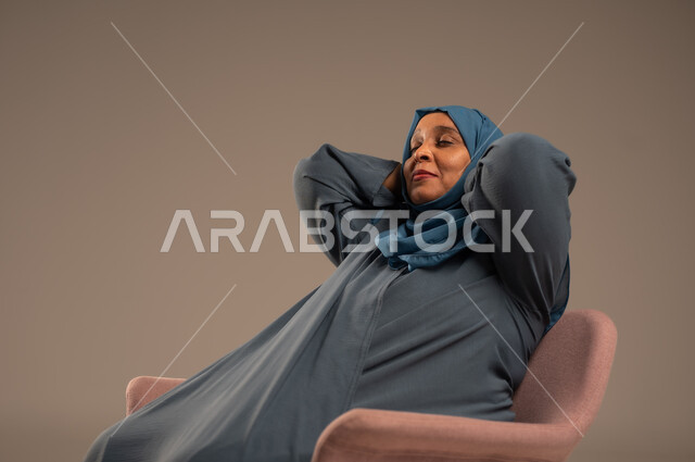 الاستمتاع بوقت الهدوء, أخذ قسط من الاسترخاء و التأمل, بورتريه لامراة مسنة عربية خليجية سعودية ترتدي الحجاب والعباءة تجلس على كرسي مريح، تعابير السرور والراحة, خلفية بيج