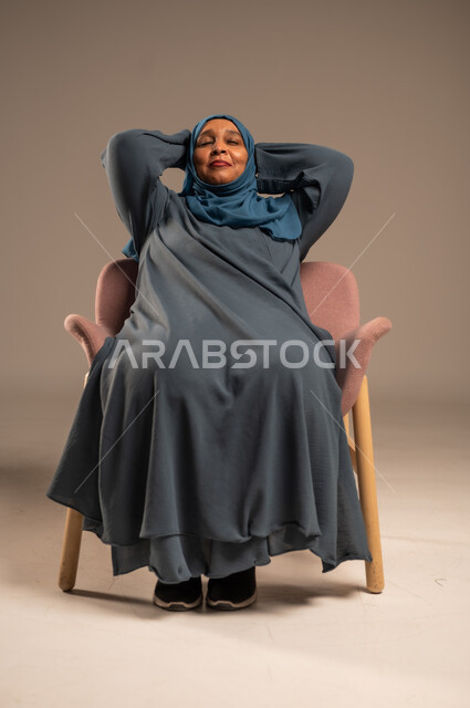 Taking a break, portrait of an elderly Saudi Arabian Gulf woman wearing a blue abaya sitting on a comfortable chair with her hands behind her head with expressions of relaxation and pleasure, gestures of meditation and enjoying quiet time, beige background