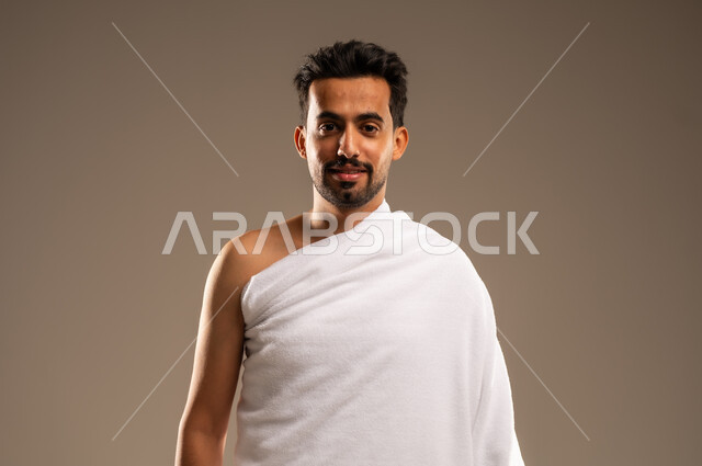 زيارة البيت الحرام في مكة المكرمة، بورتريه مقرب لرجل مسلم عربي خليجي سعودي مبتسم يرتدي لباس الاحرام ينظر الى الكاميرا بتعابير السرور, الإستعداد لأداء مناسك فريضة الحج والعمرة، الركن الخامس من أركان الاسلام، ايماءات السعادة والطمأنينة، خلفيه بيج