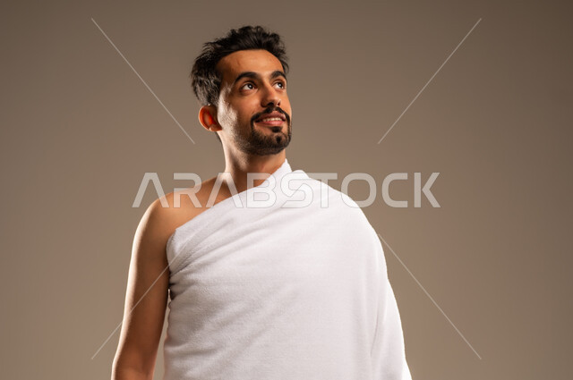 زيارة البيت الحرام في مكة المكرمة، بورتريه مقرب لرجل مسلم عربي خليجي سعودي مبتسم يرتدي لباس الاحرام ينظر الى شيء ما بتعابير السرور, الإستعداد لأداء مناسك فريضة الحج والعمرة، الركن الخامس من أركان الاسلام، ايماءات السعادة والطمأنينة، خلفيه بيج