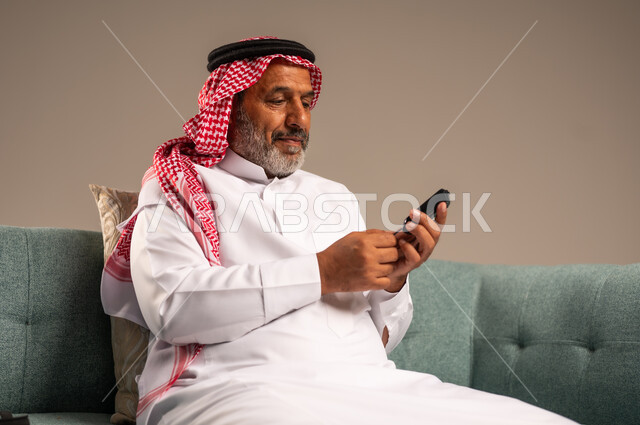 جهاز إلكتروني حديث لقياس سكر الدم لمرضى داء السكري، بورتريه مقرب لرجل مسن  عربي خليجي سعودي مريض يرتدي الشماغ والثوب التقليدي يجلس على كنبة مريحة في غرفة المعيشة يقوم بفحص نسبة السكر في دمه، الاهتمام والرعاية بالصحة، خلفية بيج