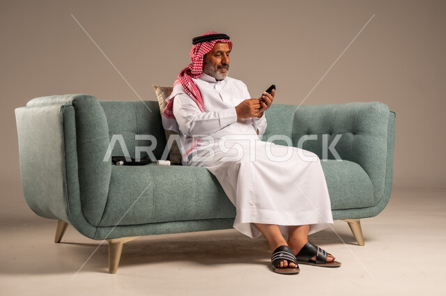جهاز إلكتروني حديث لقياس سكر الدم لمرضى داء السكري، بورتريه لرجل مسن  عربي خليجي سعودي مريض يرتدي الشماغ والثوب التقليدي يجلس على كنبة مريحة في غرفة المعيشة يقوم بفحص نسبة السكر في دمه، الاهتمام والرعاية بالصحة الجسدية، خلفية بيج