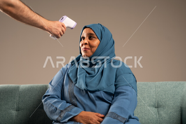 الاهتمام والرعاية بالصحة الجسدية، بورتريه لامراة مسنة عربية خليجية سعودية ترتدي الحجاب والعباءة تجلس على كرسي تقوم بمعرفة حرارة جسدها بإستخدام ميزان الحرارة الرقمي، اتباع إجراءات الوقاية والسلامة من فايروس كورونا ، خلفية بيج