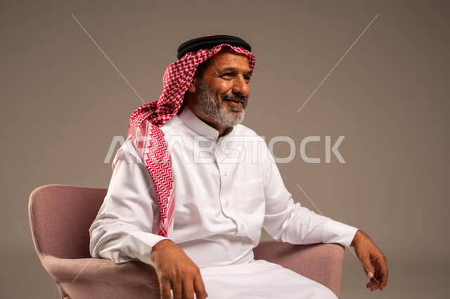 ايماءات السعادة والسرور، الاهتمام بالمظهر الخارجي، بورتريه من الجانب لرجل مسن عربي خليجي سعودي يرتدي الشماغ والثوب التقليدي يجلس على كرسي وينظر الى شيء ما، خلفية بيج
