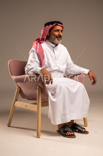 ،الاهتمام بالمظهر الخارجي، بورتريه  لرجل كبير بالسن عربي خليجي سعودي يرتدي الشماغ والثوب التقليدي يجلس على كرسي وينظر الى شيء ما بايماءات السعادة والسرور، خلفية بيج