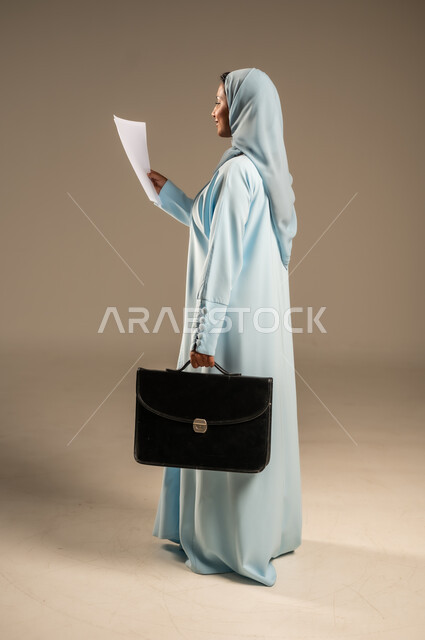 مفهوم إداة الأعمال والمشاريع، تعابير السعادة والسرور، بورتريه من الجانب لامراة عربية خليجية سعودية محجبة ترتدي عباءة ملونة تحمل بيدها مجموعه من الاوراق تنظر الى الكاميرا بايماءات الثقة بالنفس، الأناقة والاهتمام بالمظهر الخارجي، الجسم بالطول الكامل، خلفية رمادية