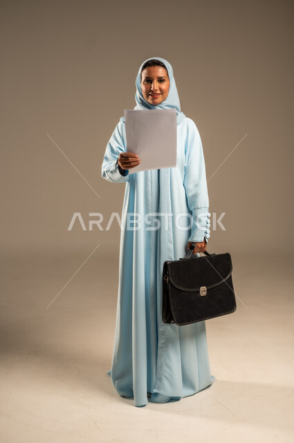 مفهوم إداة الأعمال والمشاريع، تعابير السعادة والسرور، بورتريه لامراة عربية خليجية سعودية محجبة ترتدي عباءة ملونة تحمل بيدها مجموعه من الاوراق تنظر الى الكاميرا بايماءات الثقة بالنفس، الأناقة والاهتمام بالمظهر الخارجي، الجسم بالطول الكامل، خلفية رمادية