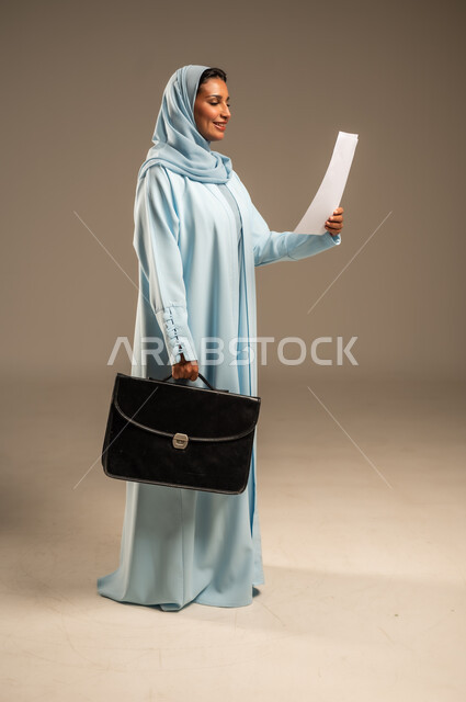 مفهوم إداة الأعمال والمشاريع، تعابير السعادة والسرور، بورتريه من الجانب لامراة عربية خليجية سعودية محجبة ترتدي عباءة ملونة تحمل بيدها حقيبة العمل الشخصية و مجموعه من الاوراق، الأناقة والاهتمام بالمظهر الخارجي، الجسم بالطول الكامل، خلفية رمادية