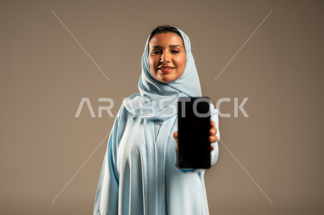Looking at the camera with gestures of happiness and pleasure, close-up portrait of a smiling Saudi Arabian Gulf woman wearing an abaya displaying a blank black screen via mobile phone, using modern and advanced devices and technologies, beige background