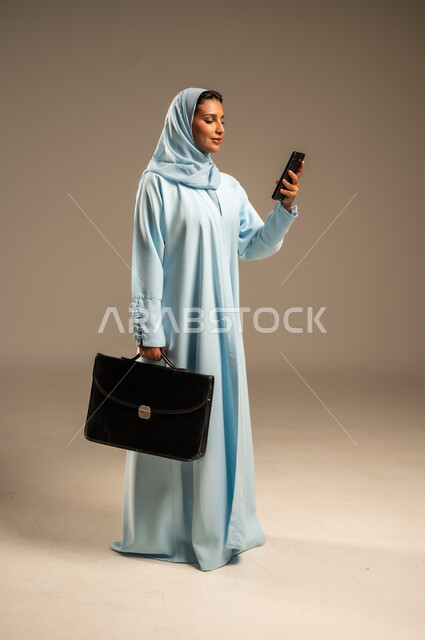 Browsing social media applications and programs, communicating with colleagues via mobile phone, portrait of a smiling Saudi Arabian Gulf woman wearing an abaya and holding a personal work bag, using modern and advanced devices and technologies, full body, beige background