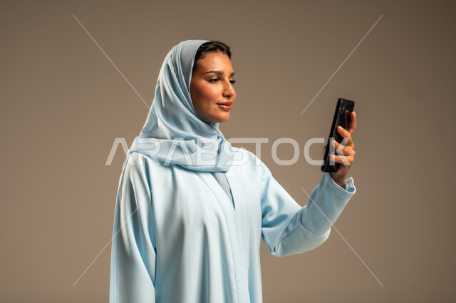تصفح التطبيقات وبرامج التواصل الاجتماعي، تبادل الأحاديث مع الأهل والأصدقاء، بورتريه لإمرأة عربية خليجية سعودية محجبة مبتسمة ترتدي العباءة تمسك بيدها الجوال، استخدام الأجهزة والتقنيات الحديثة والمتطورة، خلفية بيج