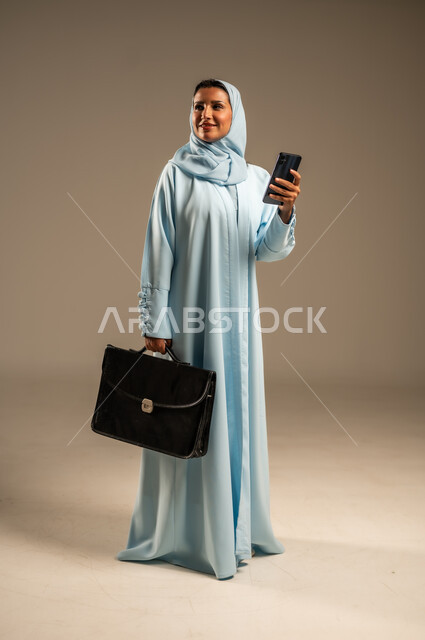 Browsing social media applications and programs, exchanging conversations with family and friends, close-up portrait of an elderly Saudi Arabian Gulf woman wearing a veil, smiling, wearing an abaya, holding a mobile phone in her hand, using modern and advanced devices and technologies, beige background