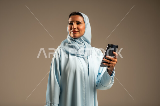تصفح التطبيقات وبرامج التواصل الاجتماعي، تبادل الأحاديث مع الأهل والأصدقاء، بورتريه لإمرأة عربية خليجية سعودية محجبة مبتسمة ترتدي العباءة تمسك بيدها الجوال، استخدام الأجهزة والتقنيات الحديثة والمتطورة، خلفية بيج